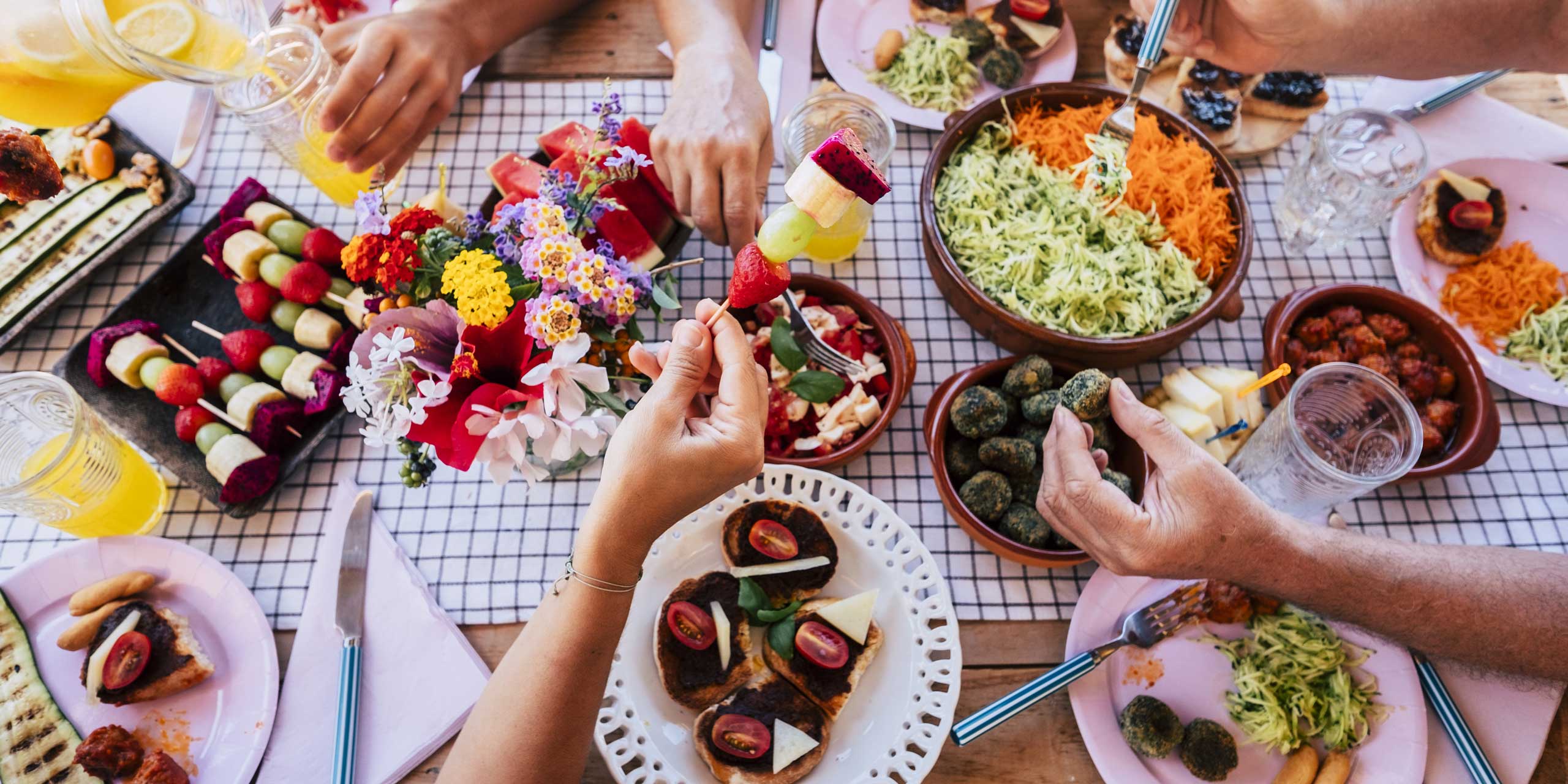 Dekorative Draufsicht einer gemeinsamen Mahlzeit mit mehreren Gerichten in Schalen, Hände greifen nach Obstspießen und Tapas