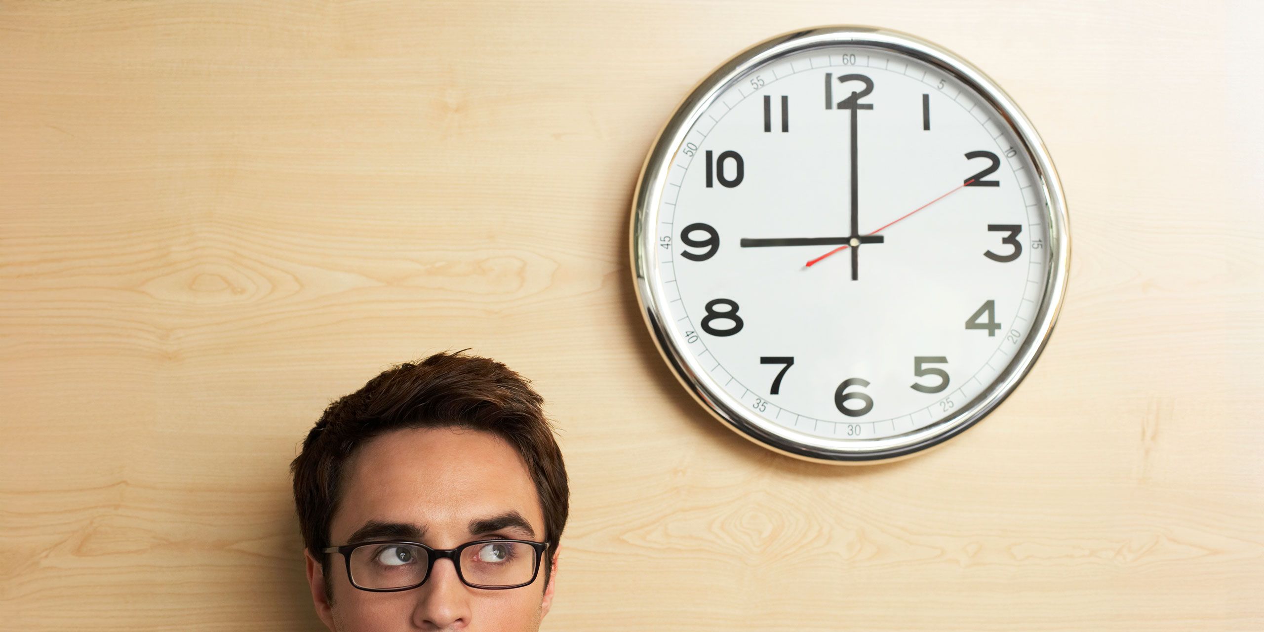 Das unten angeschnittene Gesicht eines Mannes Brille, der auf eine Wanduhr blickt. Die Uhr zeigt genau 9 Uhr.