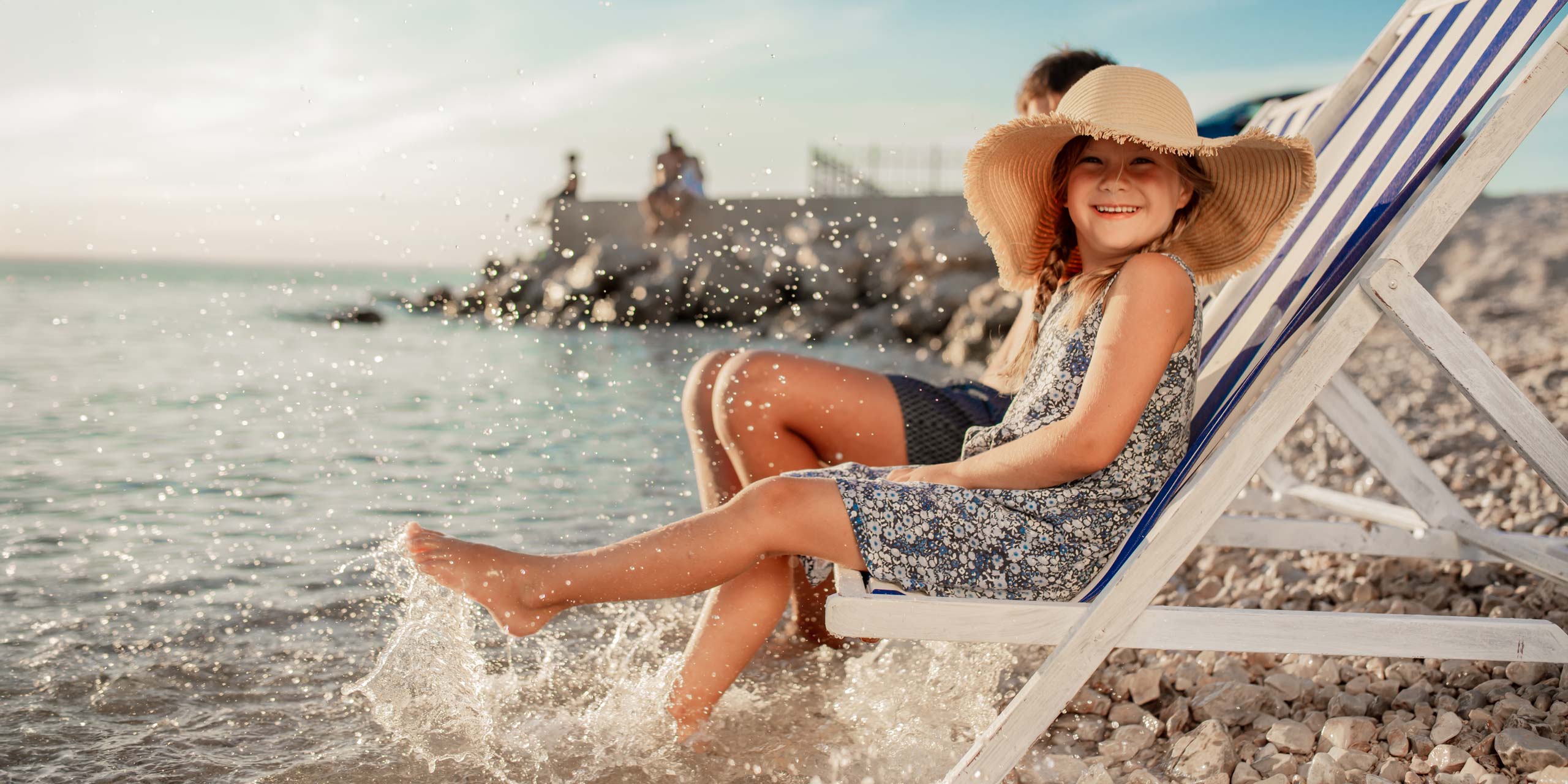 Lächelndes junges Mädchen mit Strohhut auf einem Strandstuhl auf steinigem Strand direkt am Wasser